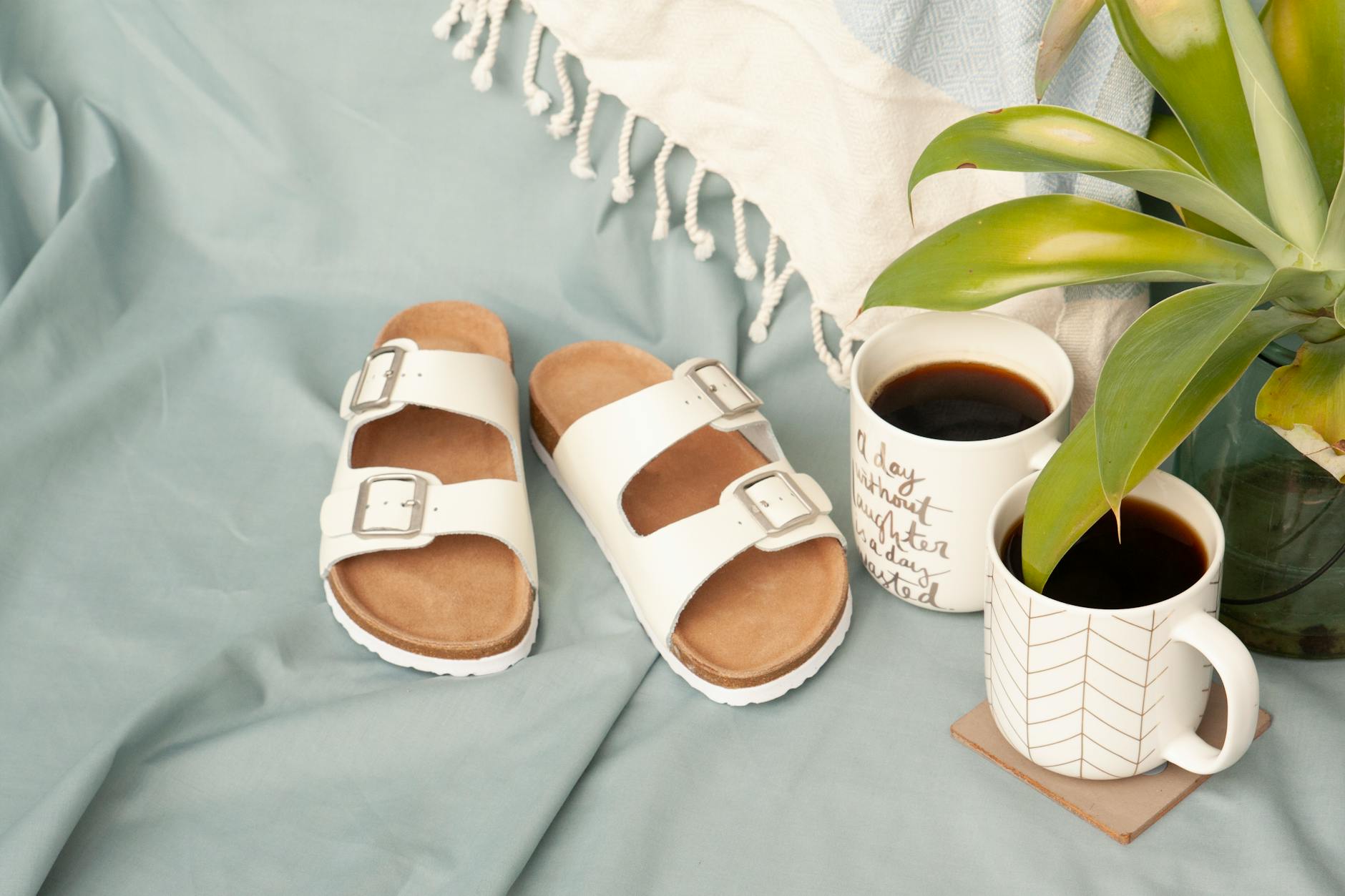 Białe sandały, kubki z kawą i roślina ułożone na podłodze