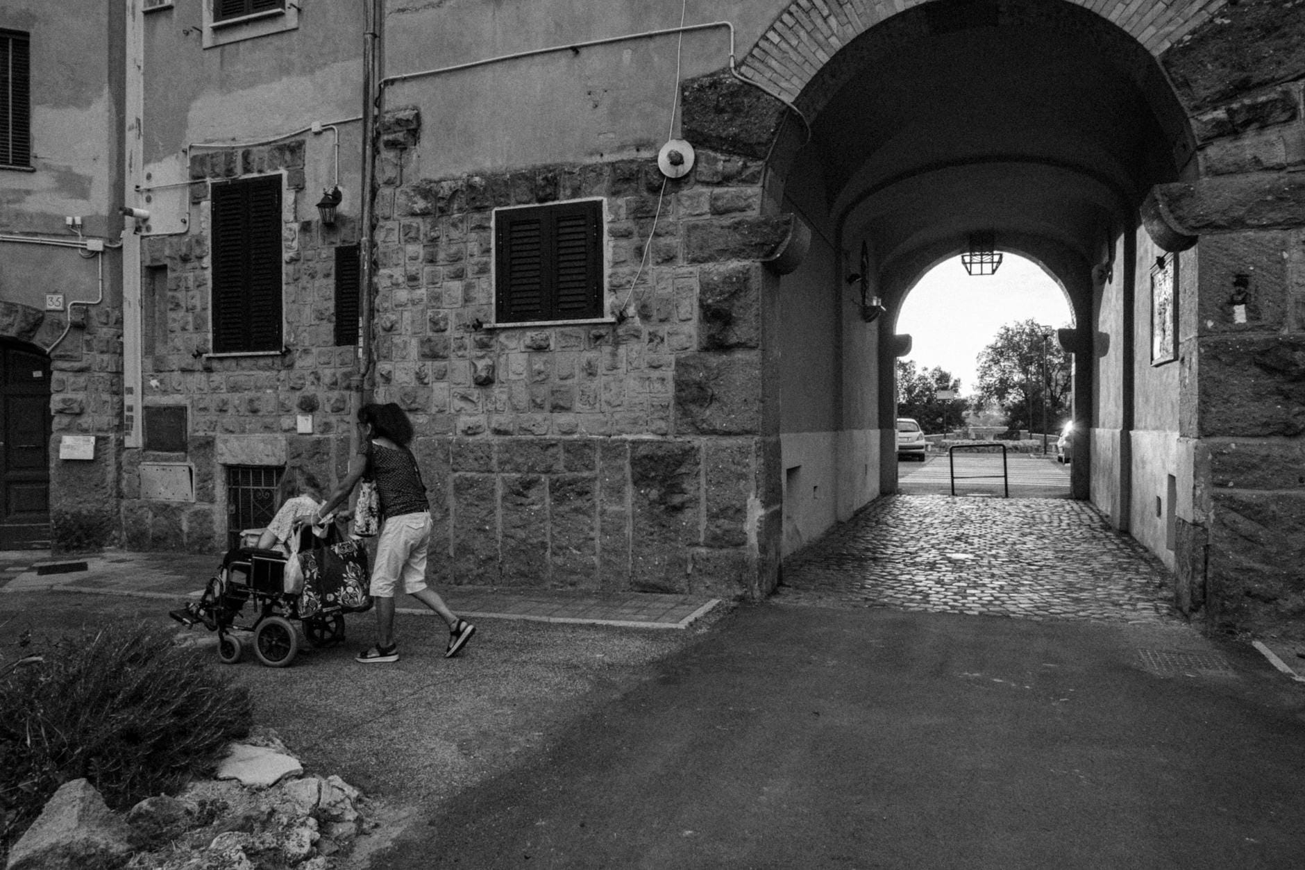 Kobieta pcha wózek inwalidzki w wąskiej, brukowanej uliczce